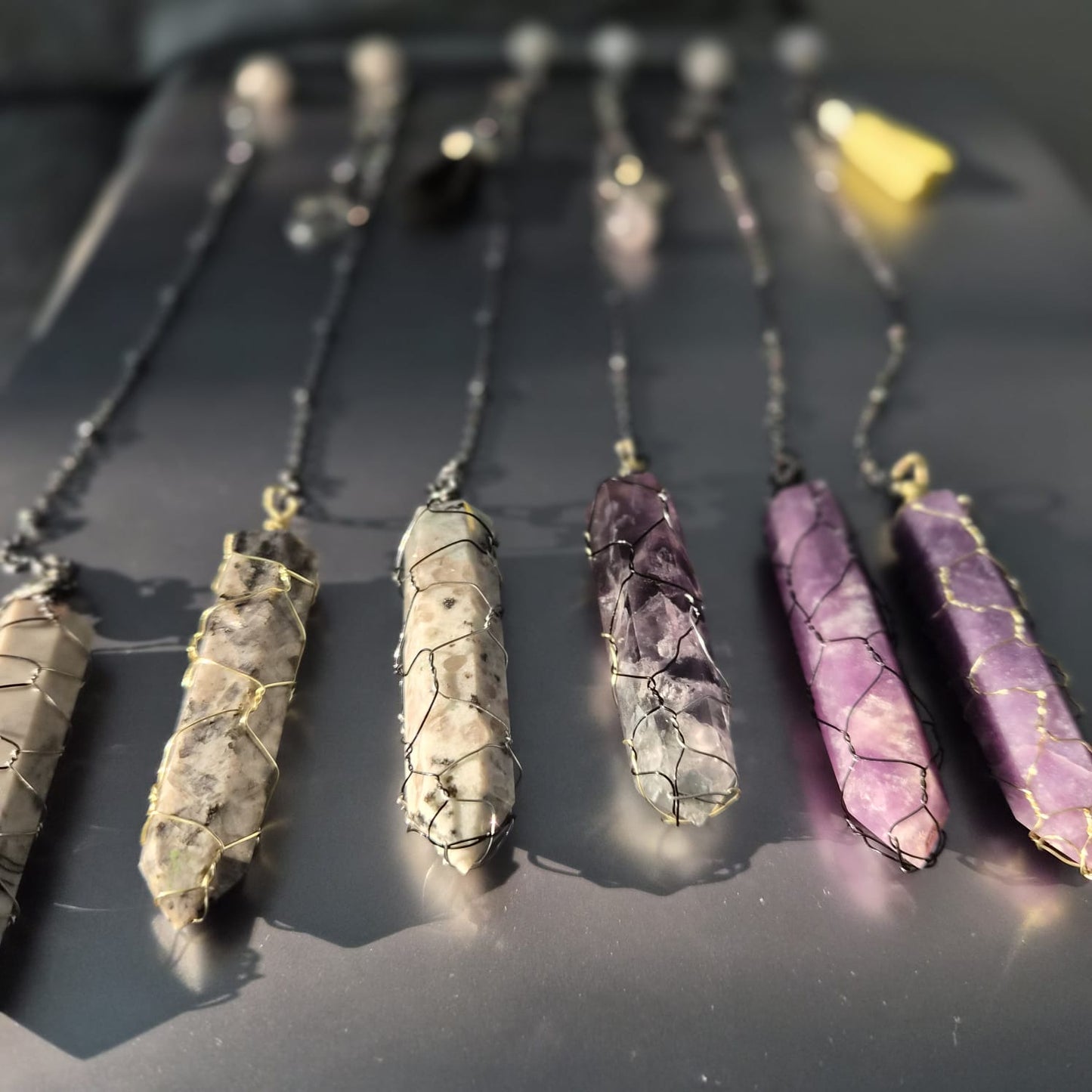 Six wire-wrapped crystal point pendulums on gunmetal chains laid on a dark grey surface — amethyst, lepidolite, rhodonite and dalmatian jasper