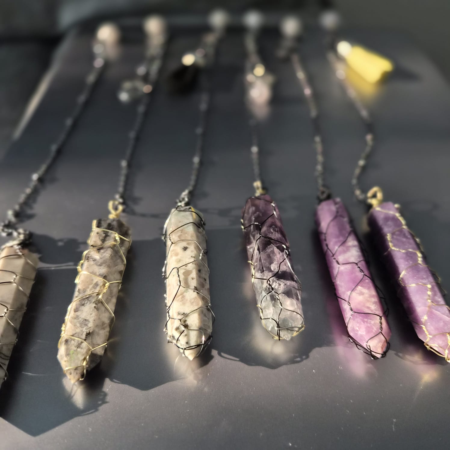 Six wire-wrapped crystal point pendulums on gunmetal chains laid on a dark grey surface — amethyst, lepidolite, rhodonite and dalmatian jasper